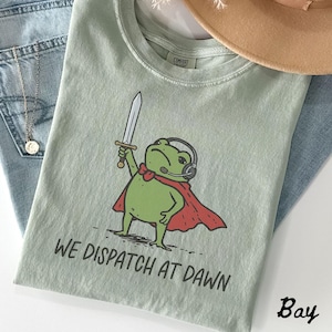May include: A sage green t-shirt featuring a graphic of a green frog wearing a red cape and black headphones while holding a sword. The text "WE DISPATCH AT DAWN" is printed in black below the frog. The shirt has a crew neckline and short sleeves.