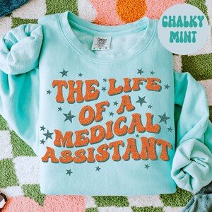 May include: A pale blue sweatshirt featuring the orange text "THE LIFE OF A MEDICAL ASSISTANT" and star graphics. The words "Chalky Mint" are in a circle. The sweatshirt is displayed on a patterned surface.