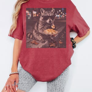 May include: A crimson short-sleeve t-shirt featuring a graphic print of a raccoon wearing black sunglasses and holding a plate of deviled eggs. The word CRIMSON is printed in the upper right corner. The shirt has a crew neckline and a relaxed fit.