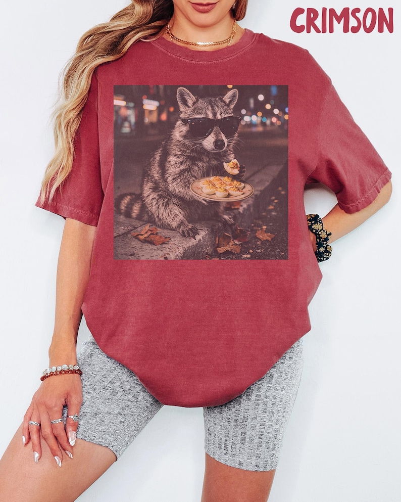 May include: A crimson short-sleeve t-shirt featuring a graphic print of a raccoon wearing black sunglasses and holding a plate of deviled eggs. The word CRIMSON is printed in the upper right corner. The shirt has a crew neckline and a relaxed fit.