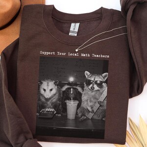 Op de afbeelding: Een donkerchocoladebruine sweater met ronde hals en een grafische print van een opossum, een stinkdier en een wasbeer. De wasbeer draagt een zonnebril en houdt een liniaal vast. De tekst boven de dieren luidt Support Your Local Math Teachers.