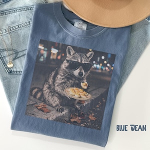 May include: A blue t-shirt featuring a graphic print of a raccoon wearing black sunglasses and holding a plate of deviled eggs. The shirt has a Comfort Colors label at the inner neckline. The text BLUE JEAN is printed in the bottom right corner.