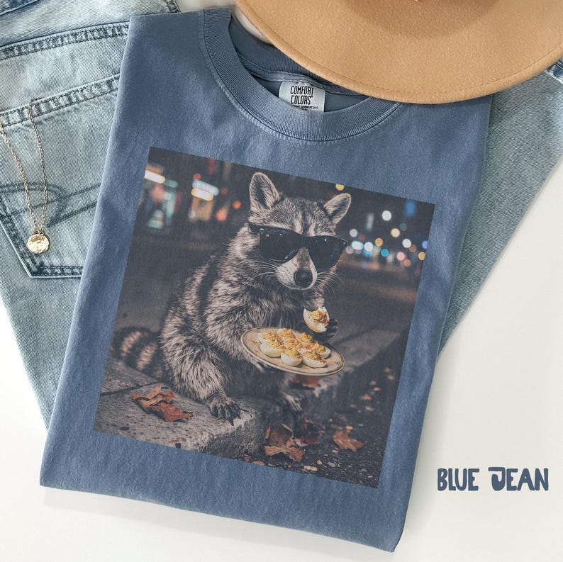 May include: A blue t-shirt featuring a graphic print of a raccoon wearing black sunglasses and holding a plate of deviled eggs. The shirt has a Comfort Colors label at the inner neckline. The text BLUE JEAN is printed in the bottom right corner.