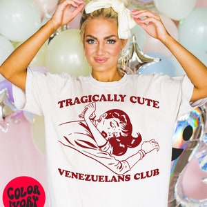 May include: White t-shirt with red text that reads "Tragically Cute" and a cartoon image of a woman with a red dress and red hair. Below the image is the text "Venezuelans Club".
