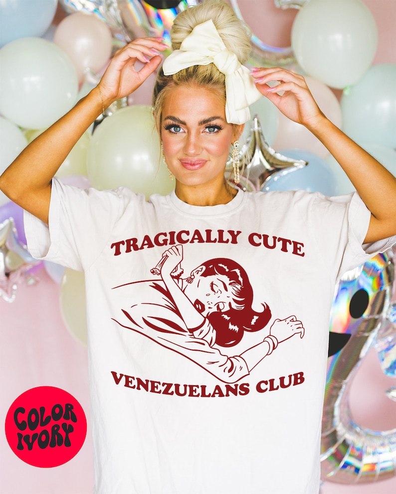 May include: White t-shirt with red text that reads "Tragically Cute" and a cartoon image of a woman with a red dress and red hair. Below the image is the text "Venezuelans Club".
