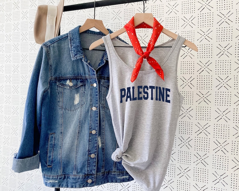 Puede incluir: Un top gris sin mangas con la palabra "PALESTINE" impresa en letras azules. El top est&aacute; colgado en una percha con una bandana roja atada alrededor del cuello.