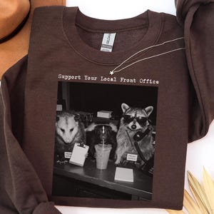 May include: A dark chocolate brown crewneck sweatshirt featuring a black and white graphic print of an opossum, a skunk, and a raccoon at an office desk. The text above the graphic reads, Support Your Local Front Office, in a typewriter font.