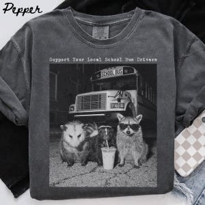 Può includere: Una t-shirt a girocollo grigio antracite con una grafica in bianco e nero di uno scuolabus, un opossum, una puzzola e un procione con occhiali da sole. Il testo sopra la grafica recita Support Your Local School Bus Drivers in carattere.
