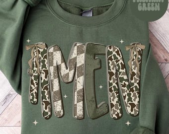 May include: Military green sweatshirt with the word "AMEN" in a distressed, textured font. The letters feature a mix of camouflage, checkered, and solid green patterns. Small star accents are scattered around the letters.