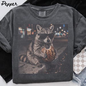 Puede incluir: Una camiseta de cuello redondo gris carbón con un estampado gráfico de un mapache con gafas de sol negras que sostiene una hogaza de pan redonda. El mapache está sobre un bordillo con un fondo de calle urbana borroso. La palabra Pepper está impresa en la esquina superior izquierda.