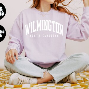 May include: A lavender sweatshirt with the text "WILMINGTON NORTH CAROLINA" in white. The model is wearing the sweatshirt and sitting with their legs crossed.