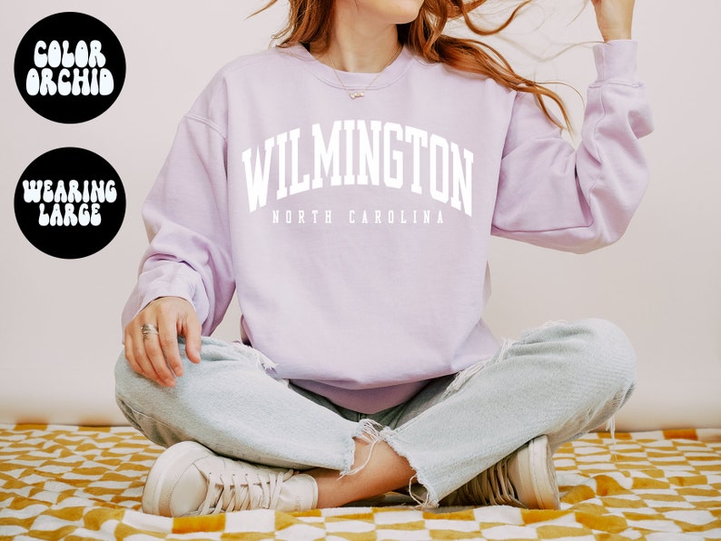 May include: A lavender sweatshirt with the text "WILMINGTON NORTH CAROLINA" in white. The model is wearing the sweatshirt and sitting with their legs crossed.