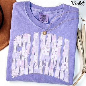 May include: A violet-colored t-shirt with the word "GRAMMA" in a patchwork design of floral and plaid patterns. A gold necklace with a pendant rests on the shirt. The shirt has a Comfort Colors tag.