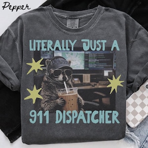 May include: A charcoal gray t-shirt featuring a graphic of a raccoon wearing a baseball cap, sunglasses, and a headset while holding a coffee cup. The text reads LITERALLY JUST A 911 DISPATCHER in light blue, accented by yellow star shapes.