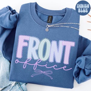 Könnte beinhalten: Indigoblaues Sweatshirt mit den Worten "FRONT office" in einem Pastell-Design. Eine silberne Halskette mit einem Sonnenblumen-Anhänger befindet sich auf dem Sweatshirt. Das Sweatshirt ist auf einer Jeans gefaltet.