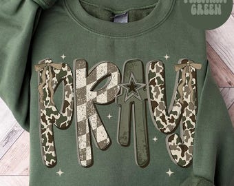 May include: Military green sweatshirt with the word "PRAY" in a unique design. The letters are a mix of camouflage, checkered, and star patterns. The text "MILITARY GREEN" is in a circle in the upper right corner.