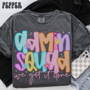 Może przedstawiać: Ciemnoszara bluza z napisem "Admin Squad" w kolorowych, bąbelkowych literach, ozdobiona kwiatami i gwiazdkami. Poniżej napis "we get it done". Widoczne etui na telefon z wzorem w szachownicę.
