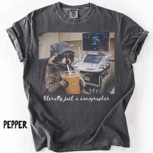 May include: Pepper-colored t-shirt featuring a raccoon wearing sunglasses and a hat, holding a drink with a straw. The image includes the text "literally just a sonographer" and a medical ultrasound machine.