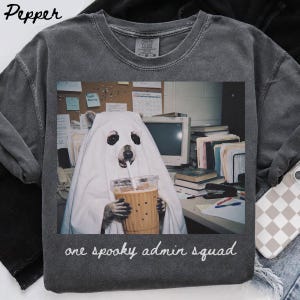 Puede incluir: Una camiseta gris carbón con un gráfico de un oso vestido con una sábana blanca de fantasma, sosteniendo un vaso de plástico con pajita. El oso está sentado en un escritorio de oficina con un ordenador y archivos. El texto dice, one spooky admin squad.