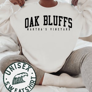 Op de afbeelding: Wit unisex sweatshirt met zwarte tekst die "Oak Bluffs Martha's Vineyard" luidt. Het sweatshirt heeft een groene golfgrafiek met de woorden "Unisex Sweatshirt" eromheen.