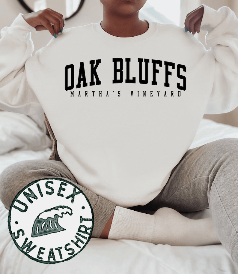 Op de afbeelding: Wit unisex sweatshirt met zwarte tekst die "Oak Bluffs Martha's Vineyard" luidt. Het sweatshirt heeft een groene golfgrafiek met de woorden "Unisex Sweatshirt" eromheen.