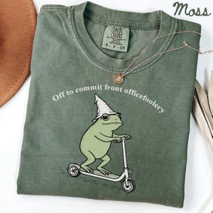 May include: Moss green t-shirt featuring a cartoon frog on a scooter, wearing a wizard hat, with the text "Off to commit front officefoolery". A gold necklace and a brown hat are also visible.