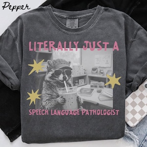 May include: A charcoal grey t-shirt featuring a black and white graphic of a raccoon wearing a baseball cap and sunglasses while holding a drink with a straw. The text reads LITERALLY JUST A SPEECH LANGUAGE PATHOLOGIST in pink with yellow stars.