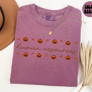 May include: A mauve-colored t-shirt with the words "human resources" in a cursive font. The shirt features a repeating pattern of orange jack-o'-lanterns and brown leaves, perfect for Halloween. A gold necklace is draped across the shirt.