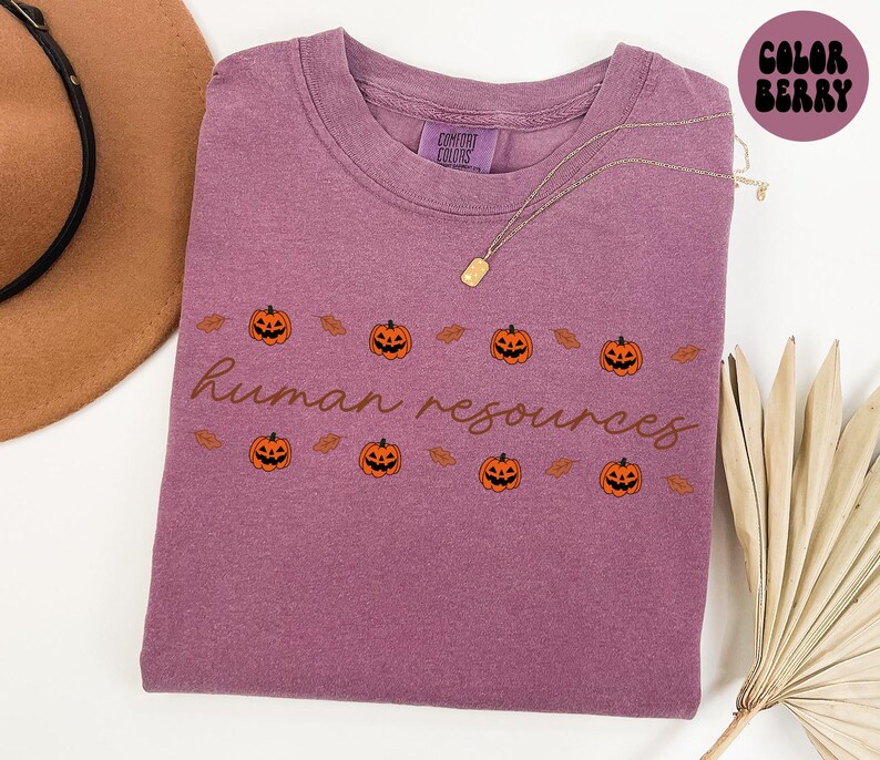 May include: A mauve-colored t-shirt with the words "human resources" in a cursive font. The shirt features a repeating pattern of orange jack-o'-lanterns and brown leaves, perfect for Halloween. A gold necklace is draped across the shirt.
