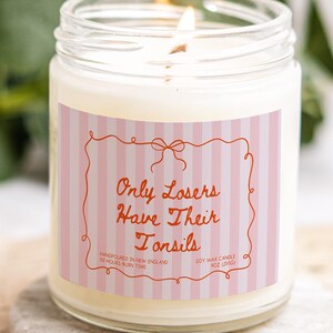 Könnte beinhalten: Eine brennende Sojawachskerze in einem durchsichtigen Glas. Das Etikett zeigt rosa und weiße Streifen mit dem Text "Only Losers Have Their Tonsils". Die Kerze ist in New England handgegossen und hat eine Brenndauer von 50 Stunden. Gewicht 255g.