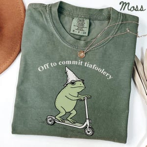 May include: Moss green t-shirt with the phrase "Off to commit tiafoolery" and a cartoon frog riding a scooter. The frog wears a wizard hat. A gold necklace and a brown hat are also visible.