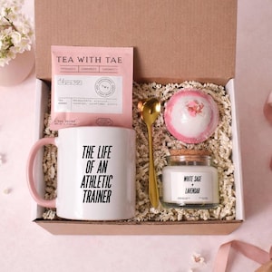 May include: Gift box with a white mug with pink handle, text "THE LIFE OF AN ATHLETIC TRAINER", pink tea packet, gold spoon, bath bomb, and a jar of White Sage + Lavendar candle.