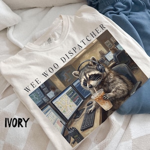 Peut inclure: Un t-shirt ivoire avec une illustration d'un raton laveur portant un casque, assis à un bureau d'ordinateur avec plusieurs écrans, tenant un café glacé, et le texte WEE WOO DISPATCHER imprimé en noir au-dessus de l'image.