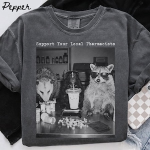 May include: A charcoal grey crewneck t-shirt featuring a black and white graphic print of an opossum, a bear, and a raccoon wearing sunglasses. The text "Support Your Local Pharmacists" is printed above the animals, which are surrounded by pill bottles.
