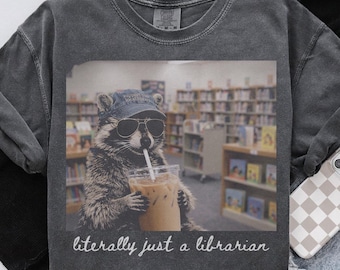 May include: A charcoal gray crew neck t-shirt featuring a graphic of a raccoon wearing a blue baseball cap and sunglasses while holding an iced coffee. The text below the graphic reads, "literally just a librarian" in a white cursive font.