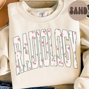 Może przedstawiać: Kremowa bluza z napisem "RADIOLOGY" w dużych, zielonych literach. Litery wypełnione są wzorem różowych truskawek. Słowo "SAND" znajduje się w brązowym okręgu w prawym górnym rogu.
