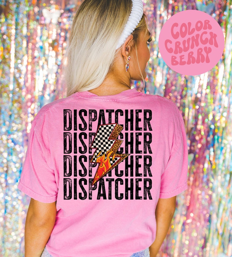 以下が含まれることがあります： ピンクのTシャツに、炎と市松模様の入った稲妻のくすんだ黒のグラフィックがプリントされています。"DISPATCHER"という文字がくすんだ黒のフォントで4回繰り返されています。