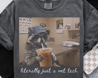 May include: A charcoal gray t-shirt featuring a graphic of a raccoon wearing a blue baseball cap and sunglasses, holding an iced coffee with a straw. Below the graphic, the text reads, "literally just a vet tech" in a white cursive font.