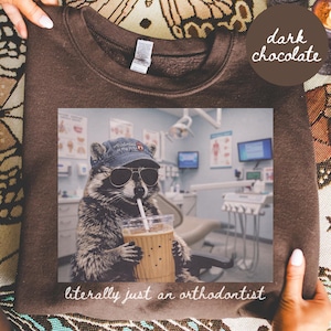 Könnte beinhalten: Ein dunkel schokoladenbraunes Sweatshirt mit einer Grafik eines Waschbären, der eine blaue Baseballkappe und eine Sonnenbrille trägt und einen Eiskaffee hält. Der Text auf der Kappe lautet 'orthodontics is my jam' und unter dem Bild 'literally just an orthodontist'.