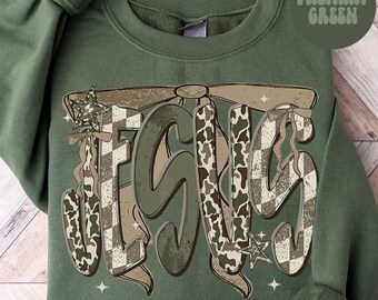 Puede incluir: Sudadera verde militar con la palabra "JESUS" en una fuente grande y estilizada. Las letras presentan una mezcla de patrones de camuflaje y cuadros, con cintas decorativas y detalles de estrellas. El texto "MILITARY GREEN" está en un círculo en la esquina superior derecha.