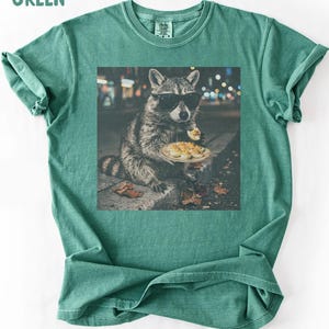 May include: A light green t-shirt featuring a graphic print of a raccoon wearing black sunglasses and eating a deviled egg from a plate. The text LIGHT GREEN is printed in the top left corner. The shirt has a crew neckline and short sleeves.