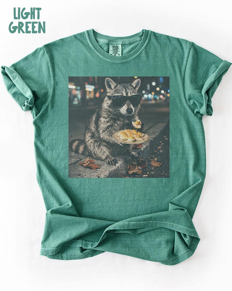 May include: A light green t-shirt featuring a graphic print of a raccoon wearing black sunglasses and eating a deviled egg from a plate. The text LIGHT GREEN is printed in the top left corner. The shirt has a crew neckline and short sleeves.