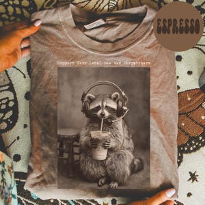 May include: A brown crew neck t-shirt featuring a graphic of a raccoon wearing a headset and holding a plastic cup with a straw. The text above the graphic reads Support Your Local Wee Woo Dispatchers. The shirt has a vintage, washed-out appearance.