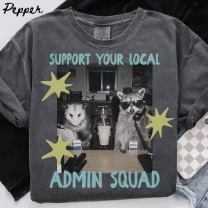 以下が含まれることがあります： ライトブルーで「SUPPORT YOUR LOCAL ADMIN SQUAD」と書かれたチャコールグレーのTシャツ。グラフィックには、デスクでサングラスとヘッドセットを着用したオポッサム、小さな哺乳類、アライグマが描かれており、4つの淡い黄色の星の形がアクセントになっています。