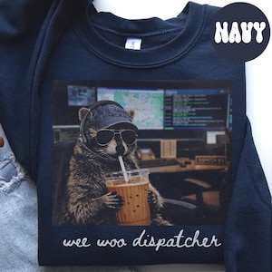 May include: A navy blue crewneck sweatshirt featuring a graphic of a raccoon wearing a baseball cap, sunglasses, and a headset while holding an iced coffee. The background shows a dispatch office with computer screens. The text reads "wee woo dispatcher".