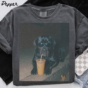 Könnte beinhalten: Ein anthrazitfarbenes T-Shirt mit Rundhalsausschnitt und einem Grafikdruck eines schwarzen Hundes mit dunkler Sonnenbrille. Der Hund ist hinter einem durchsichtigen Plastikbecher positioniert, der mit einem Eiskaffee und einem Strohhalm gefüllt ist.