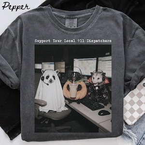 Könnte beinhalten: Ein anthrazitfarbenes T-Shirt mit Rundhalsausschnitt und einem Grafikdruck von drei kostümierten Tieren an einem Schreibtisch mit dem Text Support Your Local 911 Dispatchers. Ein Tier trägt ein Geisterlaken, eines steckt in einem Kürbis, eines trägt einen Vampirumhang.