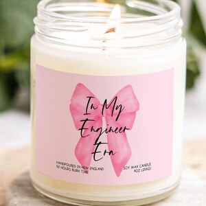 May include: A lit soy wax candle in a clear glass jar. The candle has a pink label with a watercolor bow and the text "In My Engineer Era." The label also states the candle is handpoured in New England and has a 50-hour burn time. The candle weighs 9oz (255g).