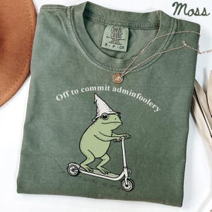 May include: Moss green t-shirt with a whimsical frog graphic riding a scooter and wearing a wizard hat. The text "Off to commit adminfoolery" is arched above the frog. A gold necklace and a brown hat are also visible.
