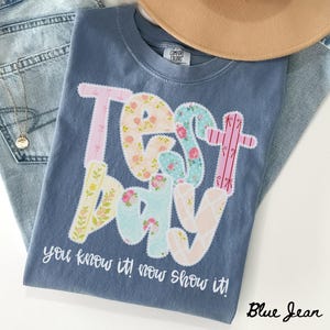 Può includere: Una maglietta blu Comfort Colors con le parole "TEST DAY" in un design patchwork floreale. Il testo "you know it! now show it!" è stampato sotto. Un cappello beige e jeans blu sono sullo sfondo.